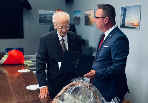 Przedstawiciel samorządu województwa składa jubilatowi życzenia, obok stół, w tle ściany pokoju.