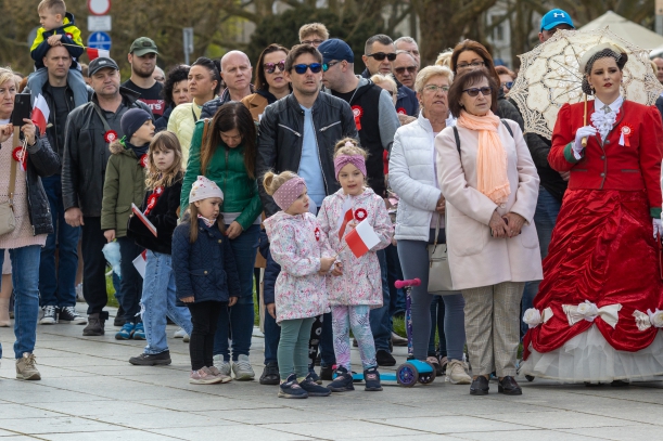 Święto Narodowe Trzeciego Maja 2023 w Szczecinie. Kilkadziesiąt osób obserwuje uroczystości przy Pomniku Czynu Polaków. W tłumie m.in. kobieta z czerwonej sukni i dziewczynki z biało-czerwonymi chorągiewkami.