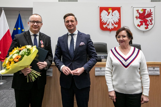 Dwaj mężczyźni w ciemnych garniturach (jeden trzyma bukiet żółtych kwiatów) i kobieta w jasnej bluzce stoją w dużej sali. W tle widać stół prezydialny, fotele, na ścienach godło państwowe i herb województwa zachodniopomorskiego.