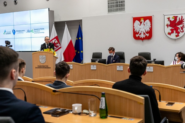 Duża sala obrad; czterech siedzących mężczyzn (zdjęcie wykonane zza ich pleców) patrzy w kierunku mównicy, przy której stoi mężczyzna w garniturze. Na mównicy - herb i napis Województwo Zachodniopomorskie. Za stołem prezydialnym siedzą mężczyzna i kobieta
