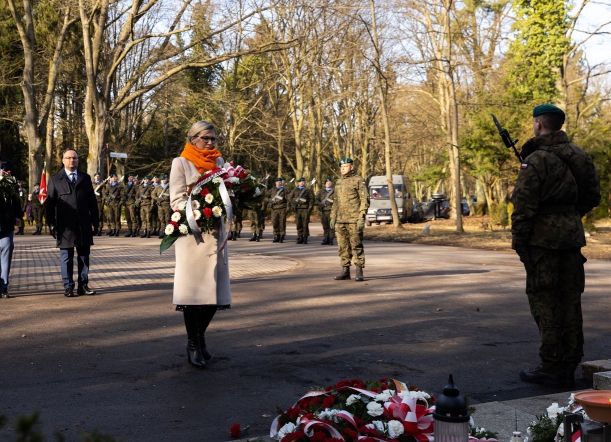 Kobieta z wiązanką kwiatów przewiązanych biało-czerwoną szarfą stoi przed pomnikiem; w tle widać kompanię żołnierzy z bronią (do salwy honorowej), przy pomniku - żołnierz pełniący asystę honorową.