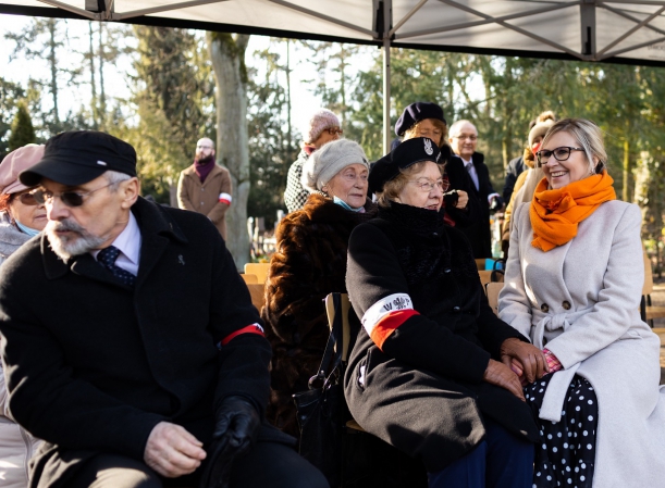 Cztery starsze osoby siedzą, kilka osób stoi, niektórzy mają biało-czerwone opaski na ramionach, obserwują uroczystość odbywająca się w plenerze; obok siedzących kombatnatów jedna młodsza kobieta w jasnym płaszczu.