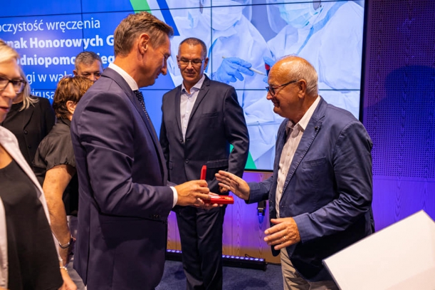 Odznaki Honorowe Gryfa Zachodniopomorskiego dla środowiska medycznego. Marszałek przypina odznakę mężczyznie dyrektorowi WSPR, w tle baner.