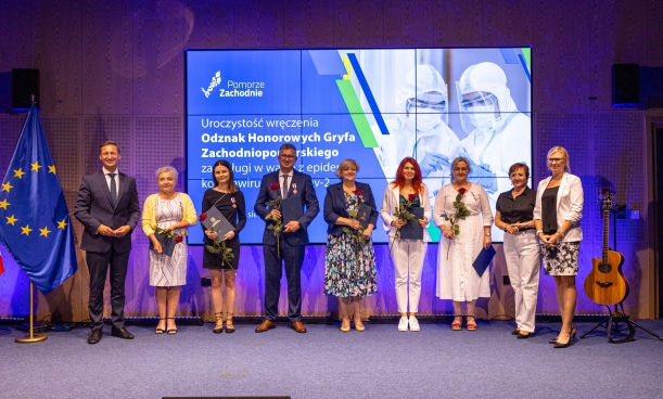 Odznaki Honorowe Gryfa Zachodniopomorskiego dla środowiska medycznego. Grupa osób wyróżniona odznakami, nagrodzeni stoją na scenie na tle baneru nawiązująćego do COViD-19
