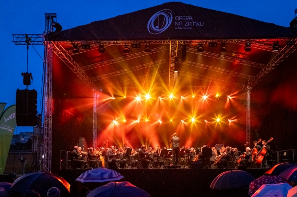 Koncert "Sierpniowe Przełomy" na placu Solidarności w Szczecinie, 27 sierpnia 2023 r. Plenerowa scena, po zmroku, oświetlona na żółto i pomarańczowo. Na scenie - muzycy i dyrygent. Przed sceną - osoby z parasolami.