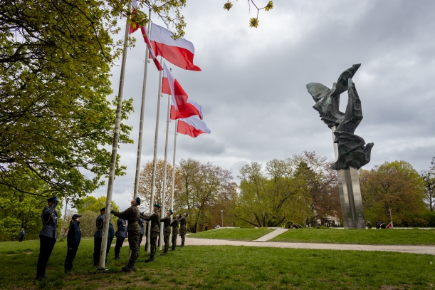 Dzień Flagi Rzeczypospolitej Polskiej. Uroczystości przy Pomniku Czynu Polaków w Szczecinie. Na pięciu masztach łopoczą biało-czerwone flagi. Przy maszatch - żółnierze, obok stoją salutujący policjanci i funkcjonariusze innych służb.