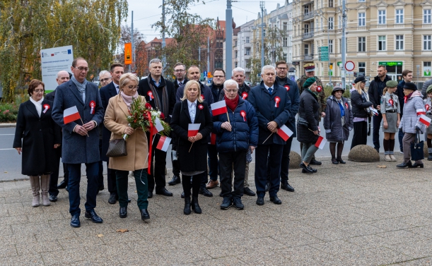 Uroczystość z okazji Narodowego Święta Niepodległości 2022 w Szczecinie. Na placu Szarych Szeregów (przy pomniku marszałka Józefa Piłsudksiego) stoi kilkadziesiąt osób z biało-czerwonymi chorągiewkami i kotylionami. Wśród nich kobieta z kwiatami.