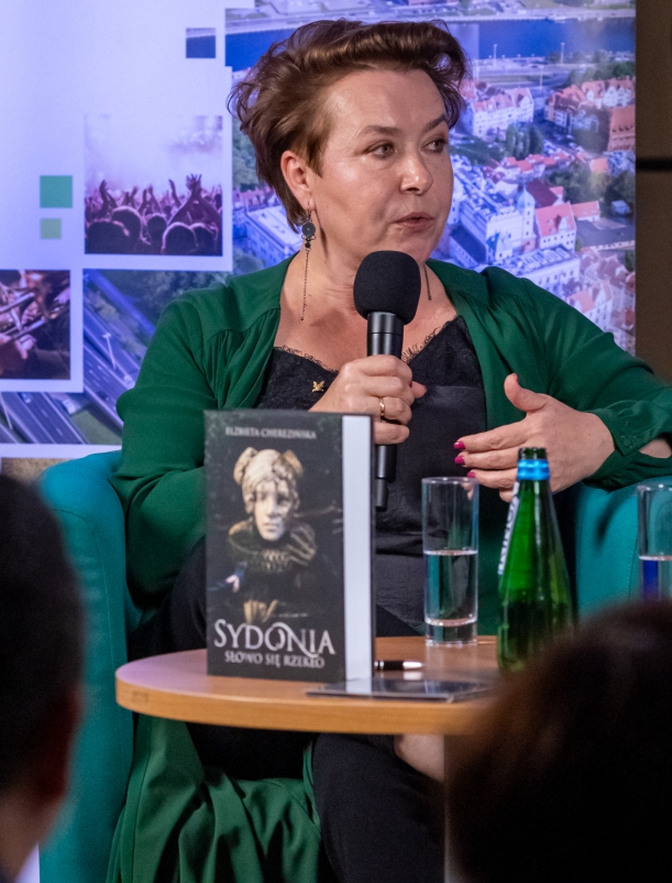 Spotkanie "Sydonia i Gryfici" (z cyklu "Otwarta społeczność") w Książnicy Pomorskiej. Na fotelu siedzi pisarka Elżbieta Cherezińsk. Trzyma mikrofon, mówi i gestykuluje. Przed nią, na stoliku - szklanka wody, butelka i książka "Sydonia. Słowo się rzekło".