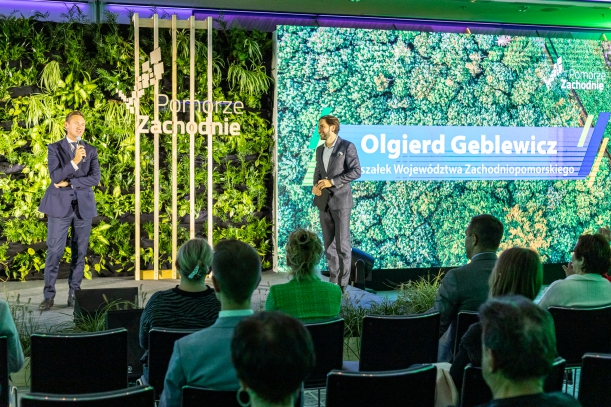 Marszałek województwa Olgierd Geblewicz w granatowym garniturze, stoi na oświetlonej scenie, z mikrofonem. Za nim ściana z roślin ozdobnych i drewniany stelaż z napisem "Pomorze Zachodnie". Obok stoi mężczyzna, a przed sceną krzesła i dziewięć osób.