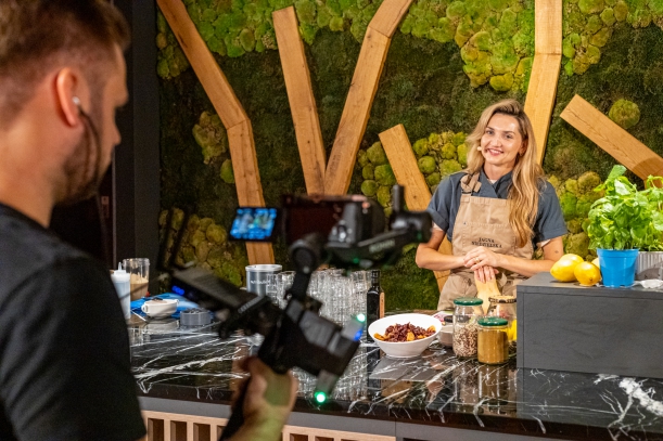 Jagna Niedzielska, propagatorka idei zero waste w gotowaniu, podczas występu w specjalnie zaaranżowanej kuchni Szczytu Ekologicznego. Stoi za dużym blatem, na którym znajdują się słoiki, półmiski, orzechy, cytryny i zioła. Przed nią stoi chłopak z kamerą.