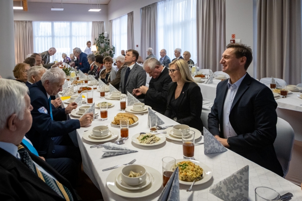 Marszałek Województwa Zachodniopomorskiego Olgierd Geblewicz oraz Członkini Zarządu Województwa Zachodniopomorskiego Anna Bańkowska siedzą za stołem podczas wigilii z kombatantami. Na długim stole biały obrus, świateczne potrawy i stroiki.