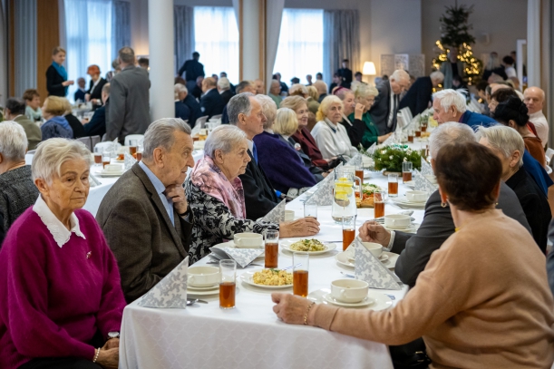 Wigilia dla kombatnatów zorganizowana przez Urząd Marszałkowski Województwa Zachodniopomorskiego w Zakładzie Aktywności Zawodowej w Dobrej. Przy długich stołach siedzi kilkadziesiat osób.