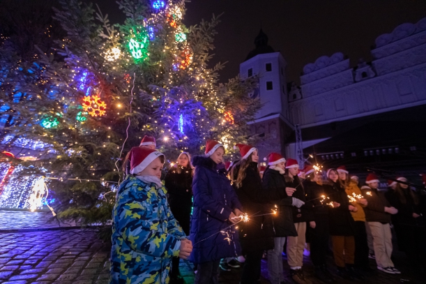 Chór śpiewający kolędy podczas włączenia oświetlenia choinki, w tle choinka.
