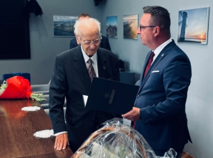 Przedstawiciel samorządu województwa składa jubilatowi życzenia, obok stół, w tle ściany pokoju.