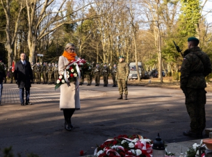 Kobieta z wiązanką kwiatów przewiązanych biało-czerwoną szarfą stoi przed pomnikiem; w tle widać kompanię żołnierzy z bronią (do salwy honorowej), przy pomniku - żołnierz pełniący asystę honorową.