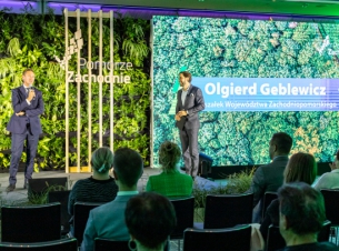 Marszałek województwa Olgierd Geblewicz w granatowym garniturze, stoi na oświetlonej scenie, z mikrofonem. Za nim ściana z roślin ozdobnych i drewniany stelaż z napisem "Pomorze Zachodnie". Obok stoi mężczyzna, a przed sceną krzesła i dziewięć osób.